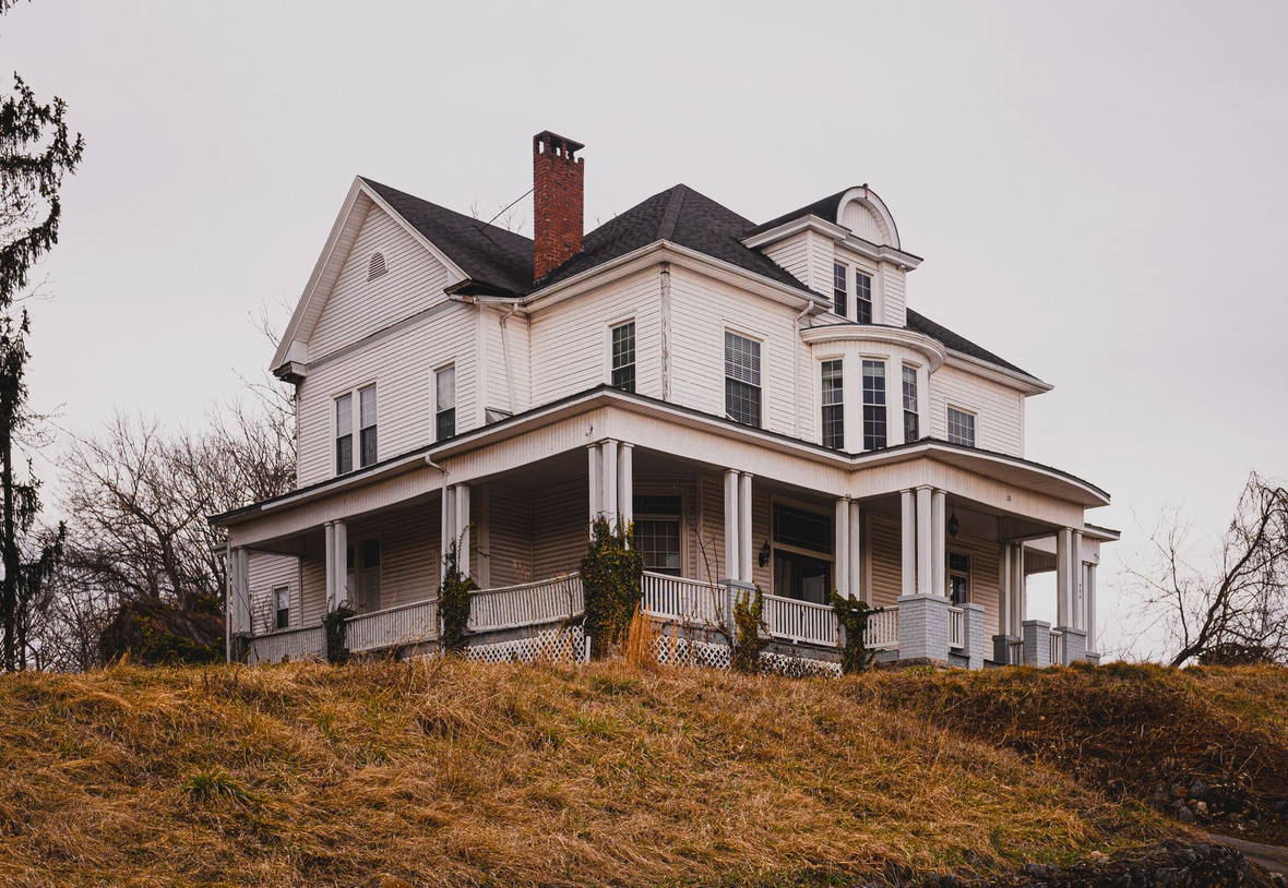 Licensed stock photograph: a tall old white clapboard house with a wraparound porch, a dark shingle roof and a brick chimney, under an overcast sky.