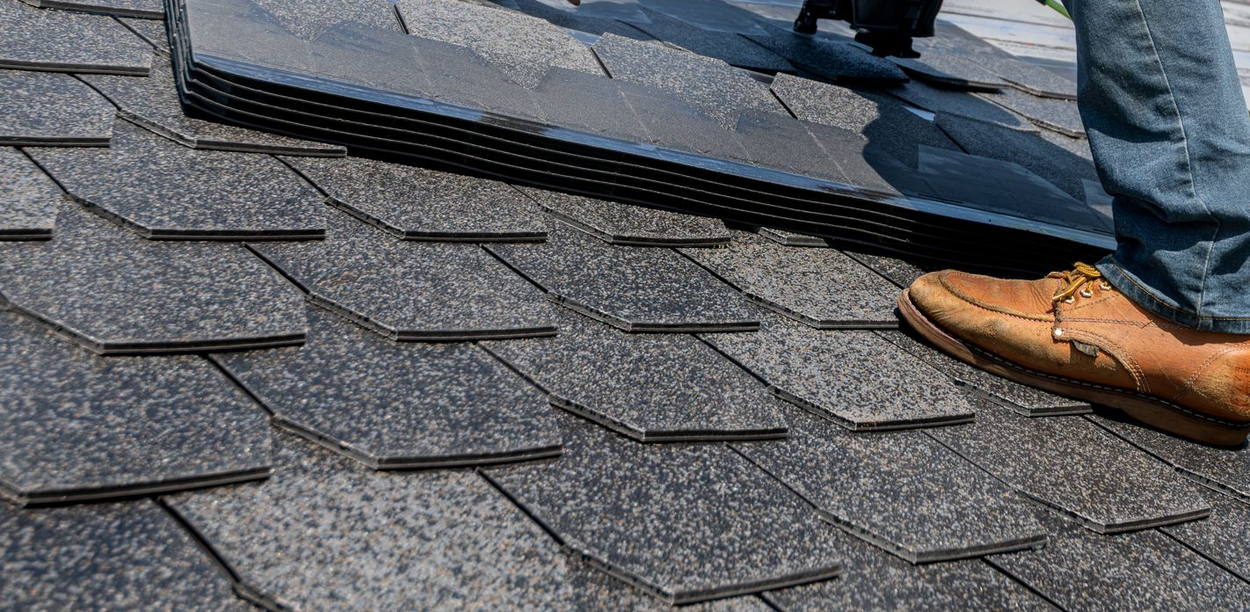Licensed stock photograph: a course of asphalt shingles being set on a roof, with a nail gun and a work boot on the finished courses below.